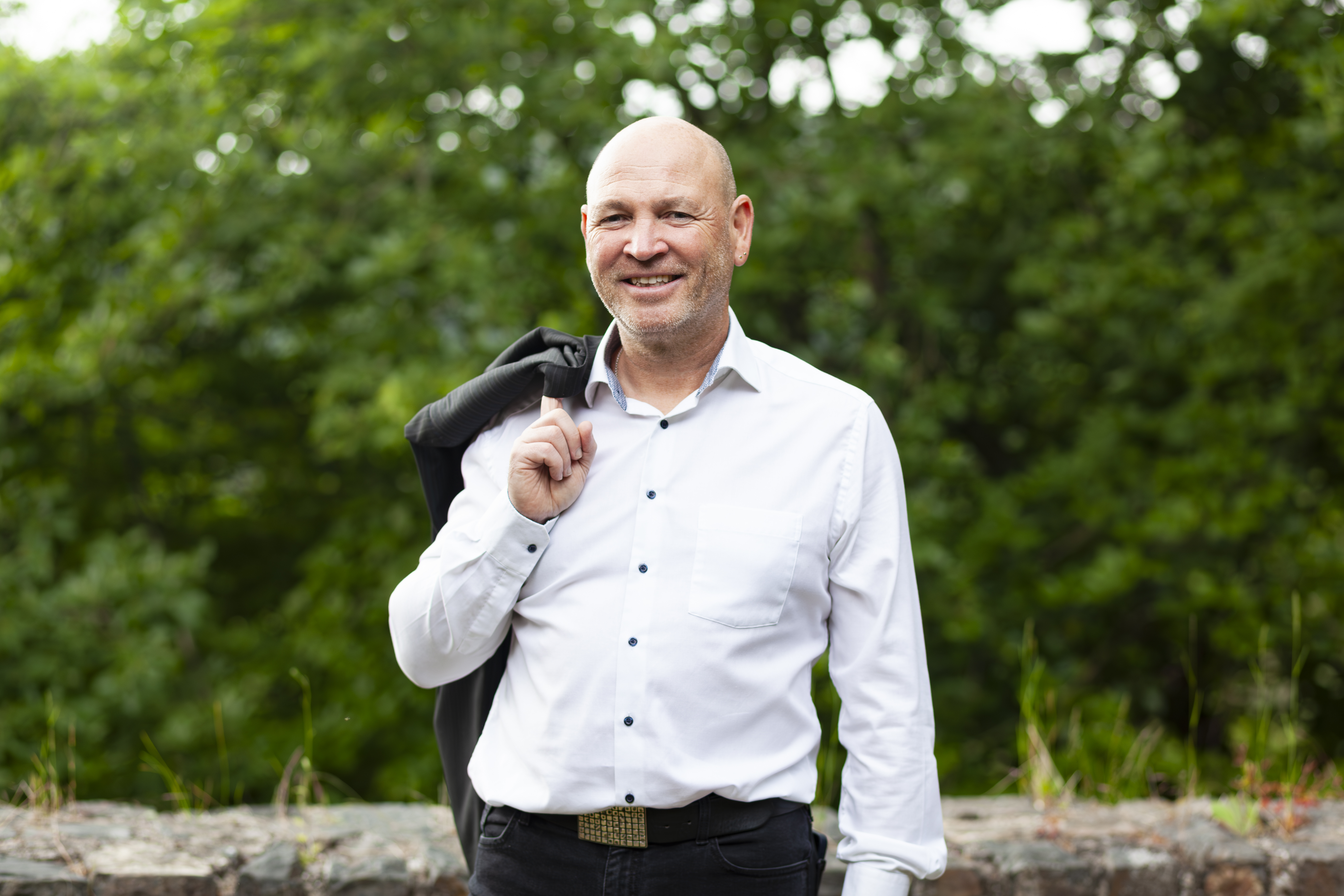 ein weisser Mann mit Glatze und weissem Hemd, das Jackett ueber der Schulter, im Hintergrund Landschaft