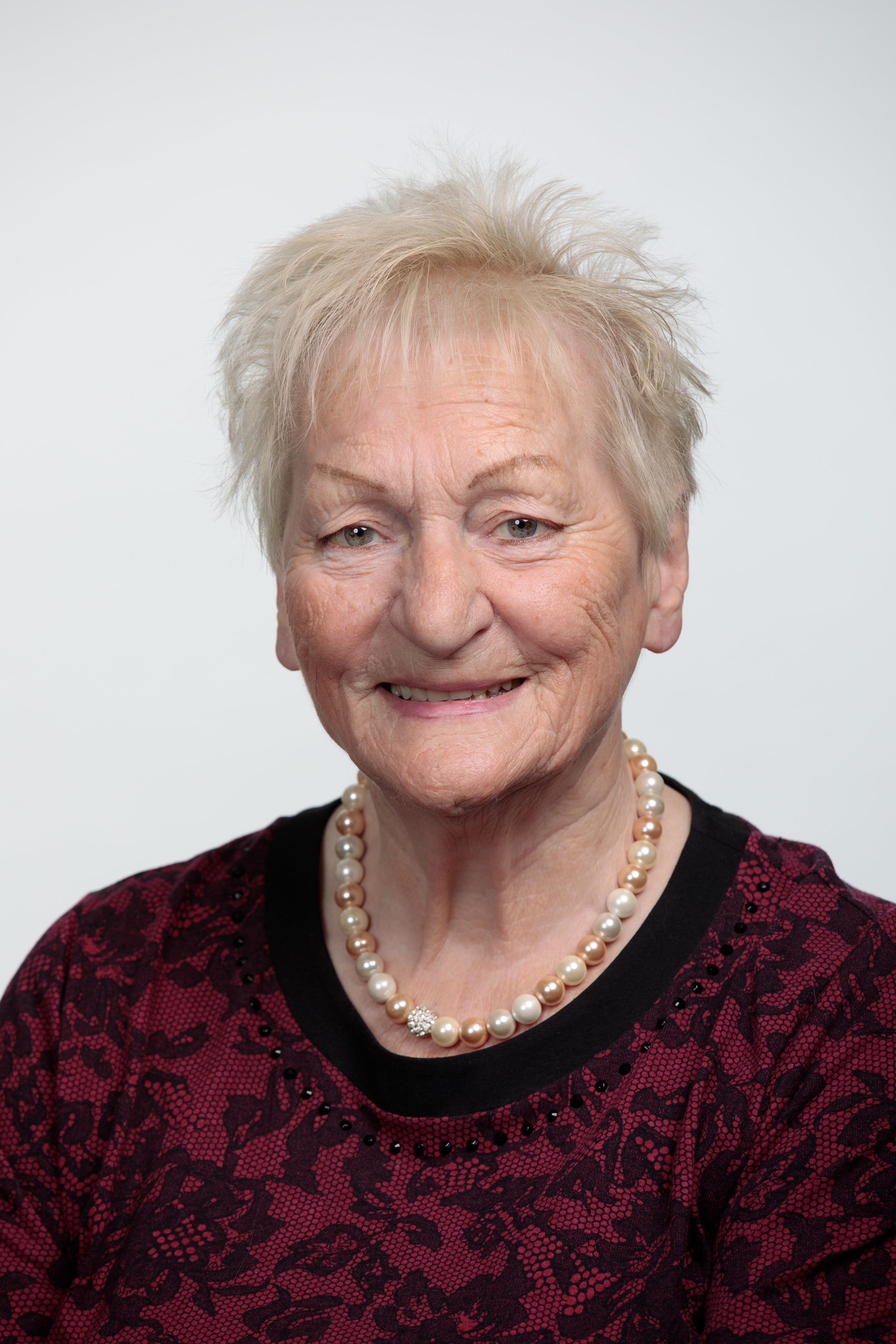 Portraitbild einer aelteren blonden weisse Frau mit kurzen Haaren mit einer Perlenkette und einem schwarz-roten Pullover