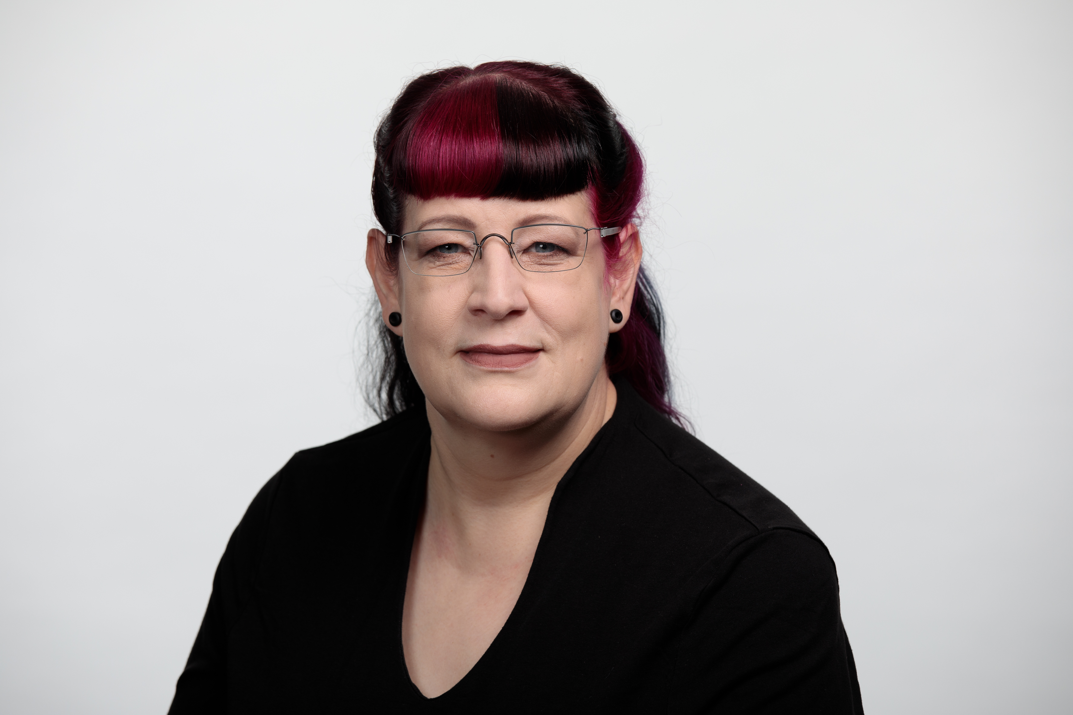 Portraitbild einer weissen Frau mit schwarzer Kleidung und Brille, dunklen langen Haaren und schwarz-violett gefaerbtem Pony